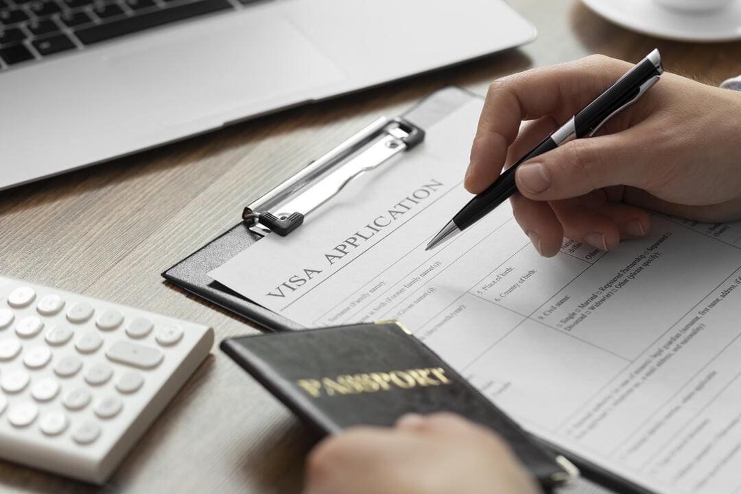 A imagem apresenta um formulário de aplicação de visto, uma mão preenchendo com uma caneta preta e a outra mão segurando um passaporte.