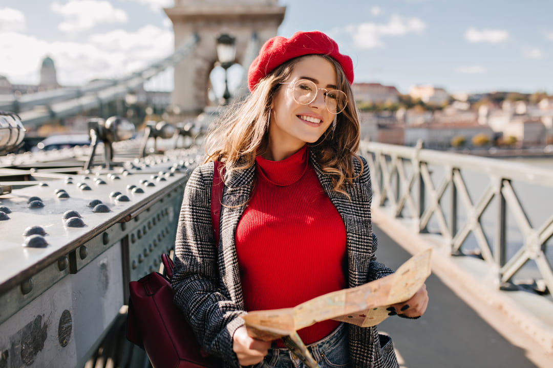 Um jovem mulher, com boina vermelha, blusa de gola alta também vermelha e casaco cinza, segura um mapa nas mãos. Ao fundo da imagem, vemos uma ponte e uma cidade medieval.